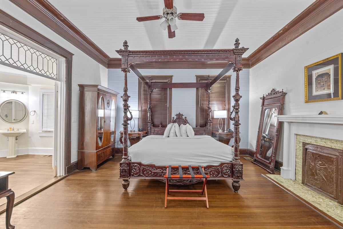 A four-poster bed with a wooden frame and white bedding, a wooden bench at the foot of the bed, and a ceiling fan above.
