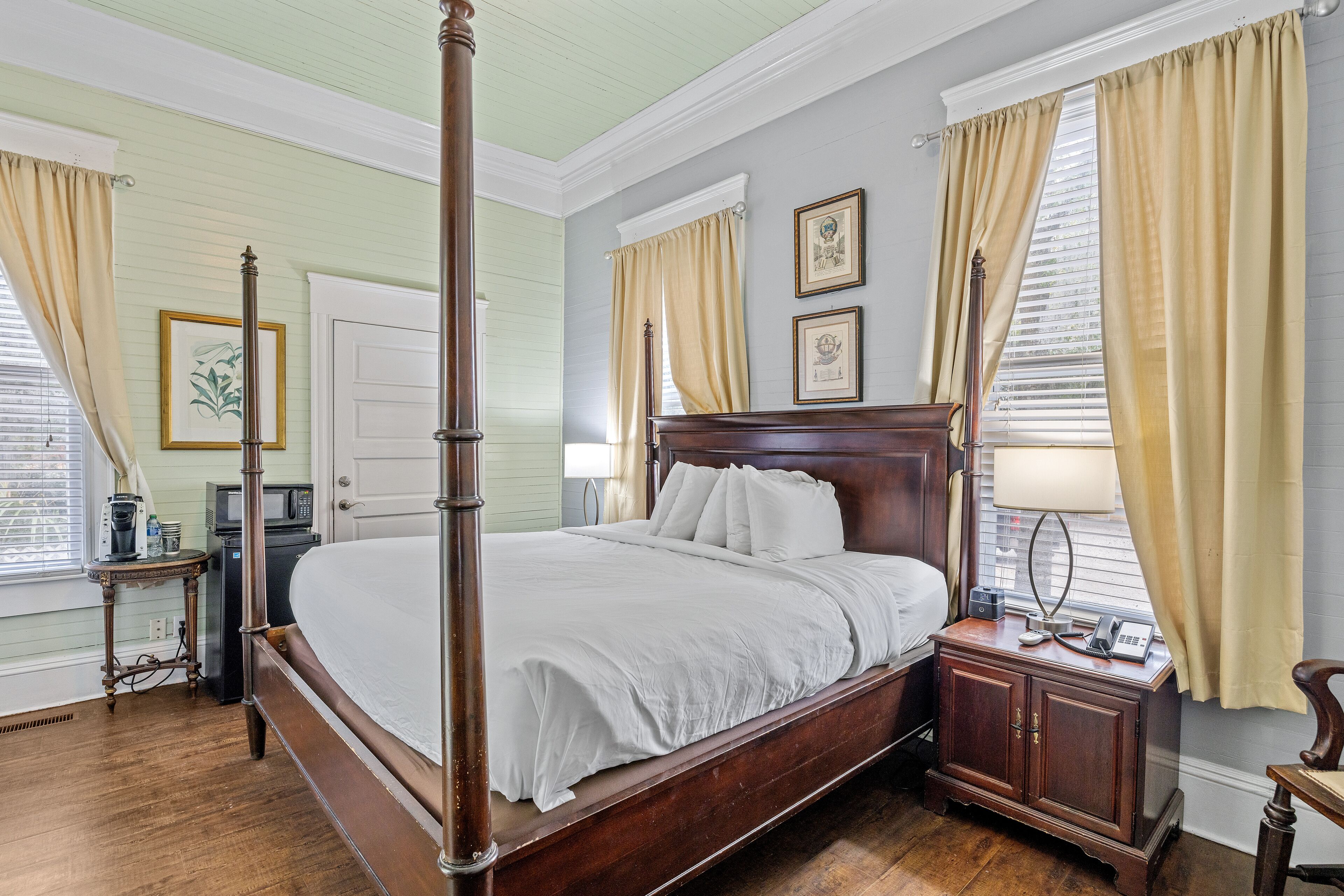 Een kamer met een hemelbed, houten vloer, een witte deur en een kleine tafel met een lamp.