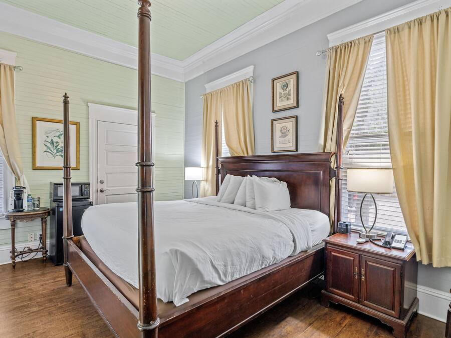 A four-poster bed in a room with a wooden floor, a white door, and a small table with a lamp.