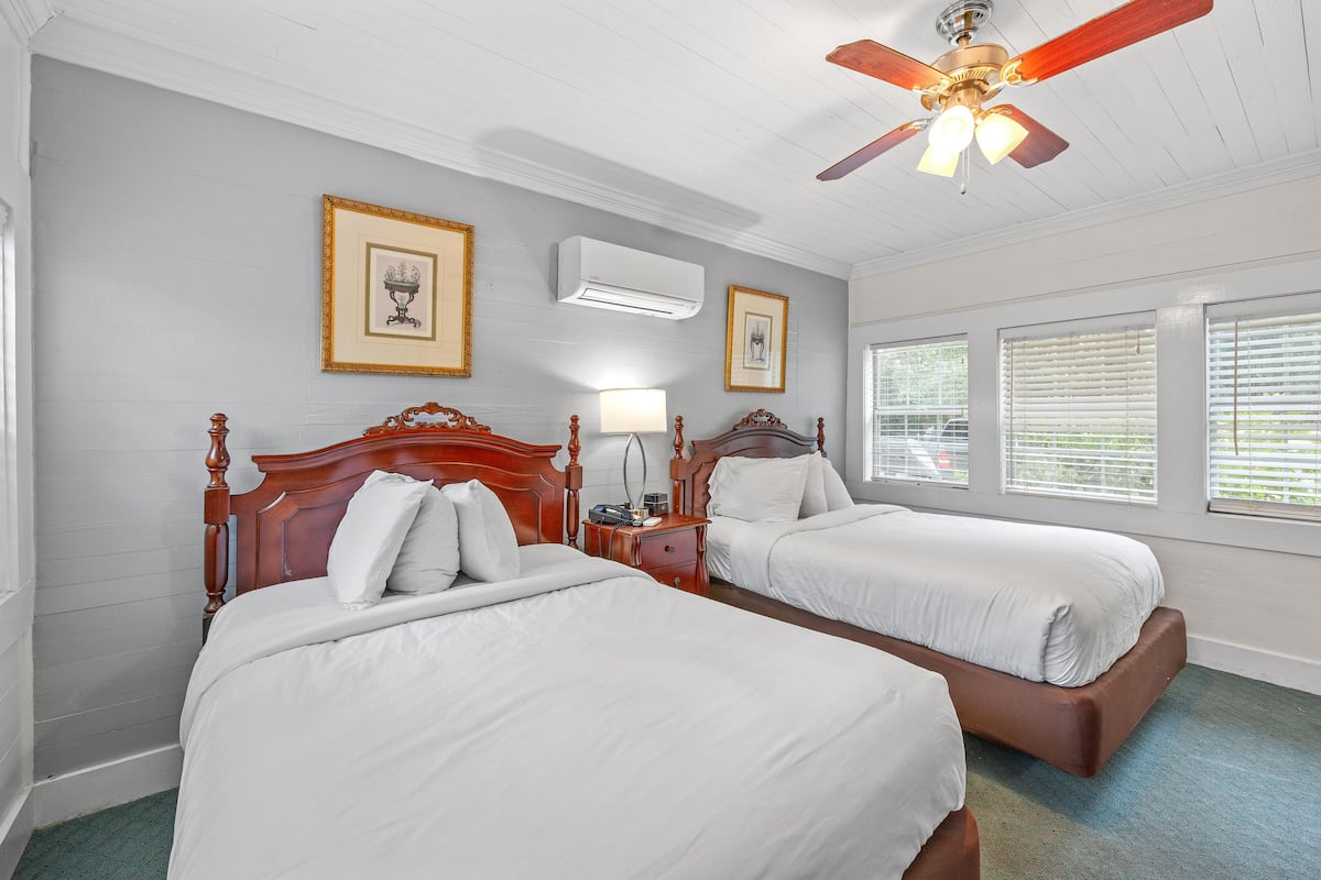 A hotel room with two beds, wooden headboards, a ceiling fan, and framed artwork on the walls.