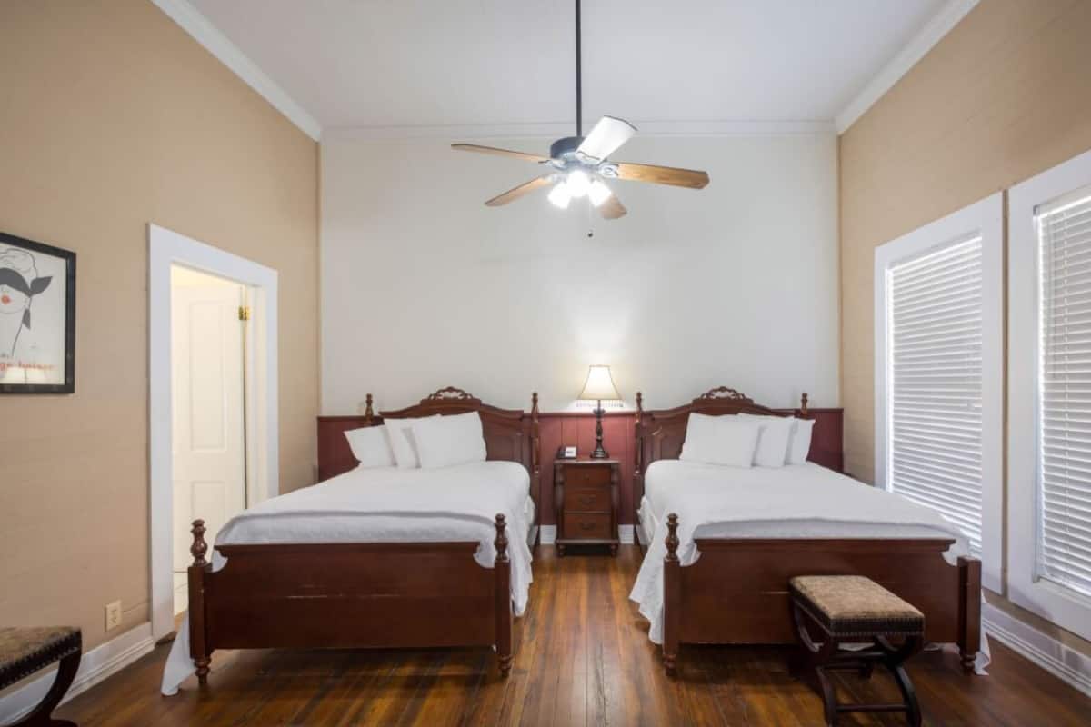 A double room with two beds, a ceiling fan, a nightstand with a lamp, and a window with blinds.