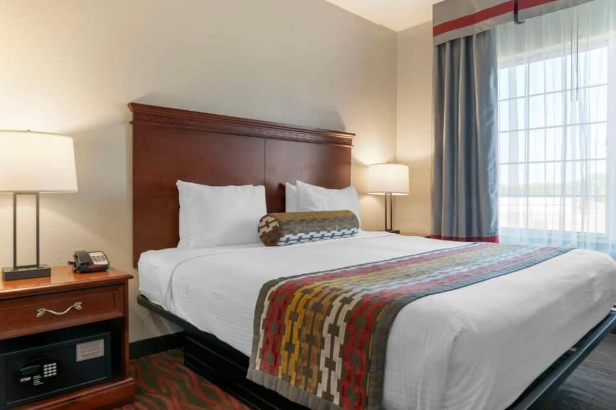 Habitación de hotel con cabecera de madera, cama con ropa blanca, dos lámparas de noche, mesita de noche con teléfono y caja fuerte, y ventana con cortinas.