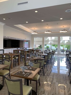 Un restaurant spacieux avec de hauts plafonds, un éclairage moderne suspendu et de grandes fenêtres offrant une vue sur l’extérieur.