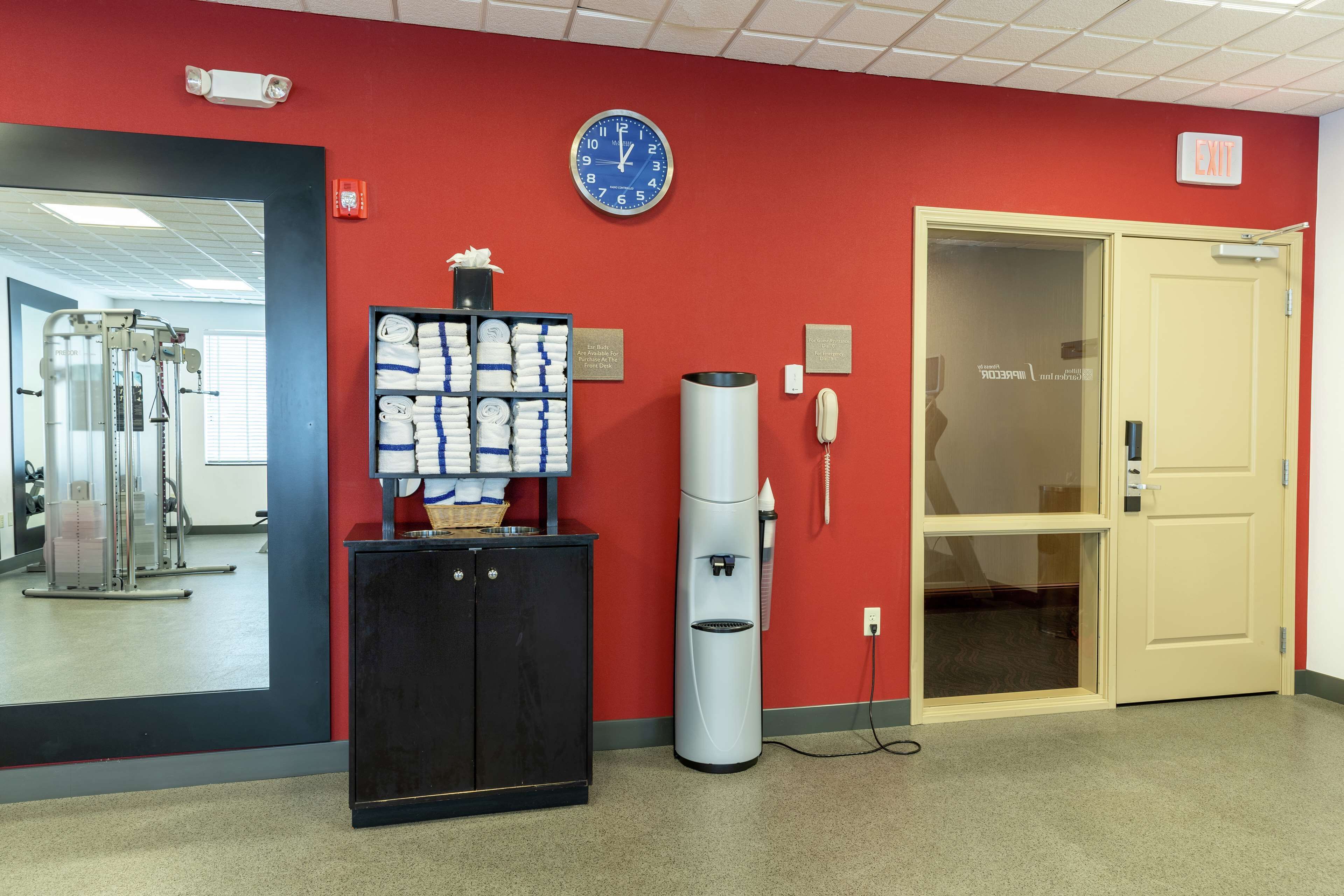 Une salle de sport équipée de matériel d’exercice, d’un distributeur d’eau, d’une horloge et d’une porte avec un panneau indiquant la sortie.