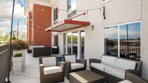 Un patio exterior con muebles de mimbre, un toldo rojo y vistas de la ciudad a través de grandes ventanales.