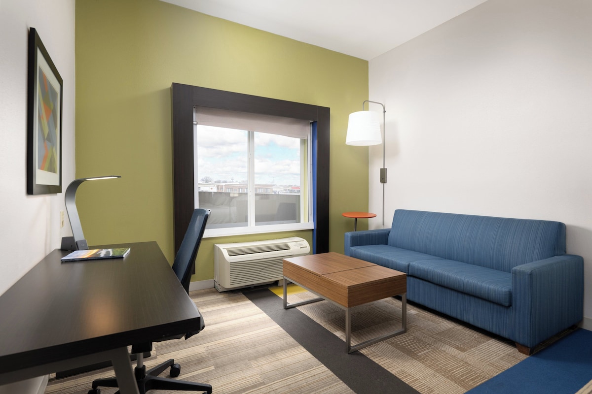 Une chambre moderne équipée d’un bureau, d’un canapé bleu, d’une lampe et d’une fenêtre donnant sur la ville.