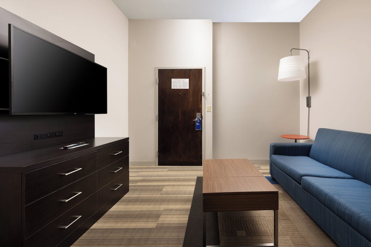 Ein modernes Hotelzimmer mit Flachbildfernseher, einem hölzernen Schminktisch, einem blauen Sofa, einem kleinen Tisch und einer Stehlampe.