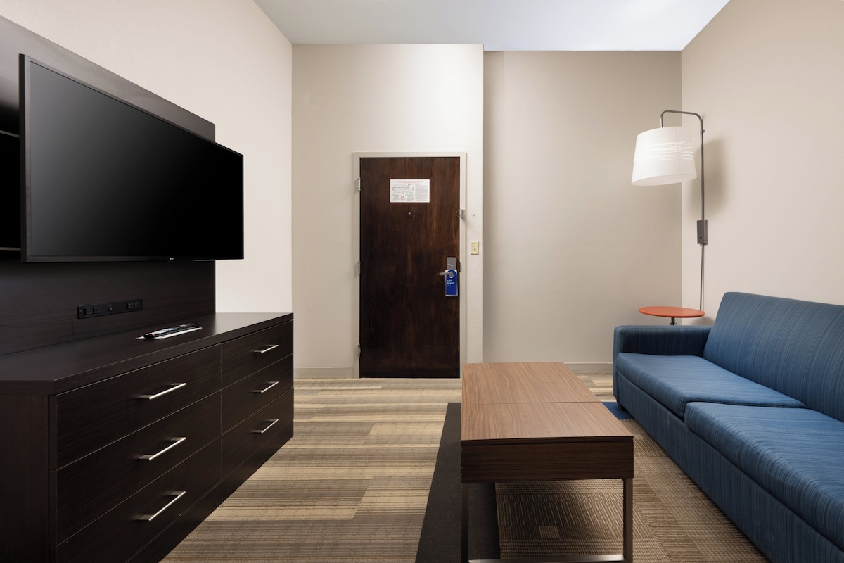 Une chambre d’hôtel moderne équipée d’un téléviseur à écran plat, d’une commode en bois, d’un canapé bleu, d’une petite table et d’un lampadaire.