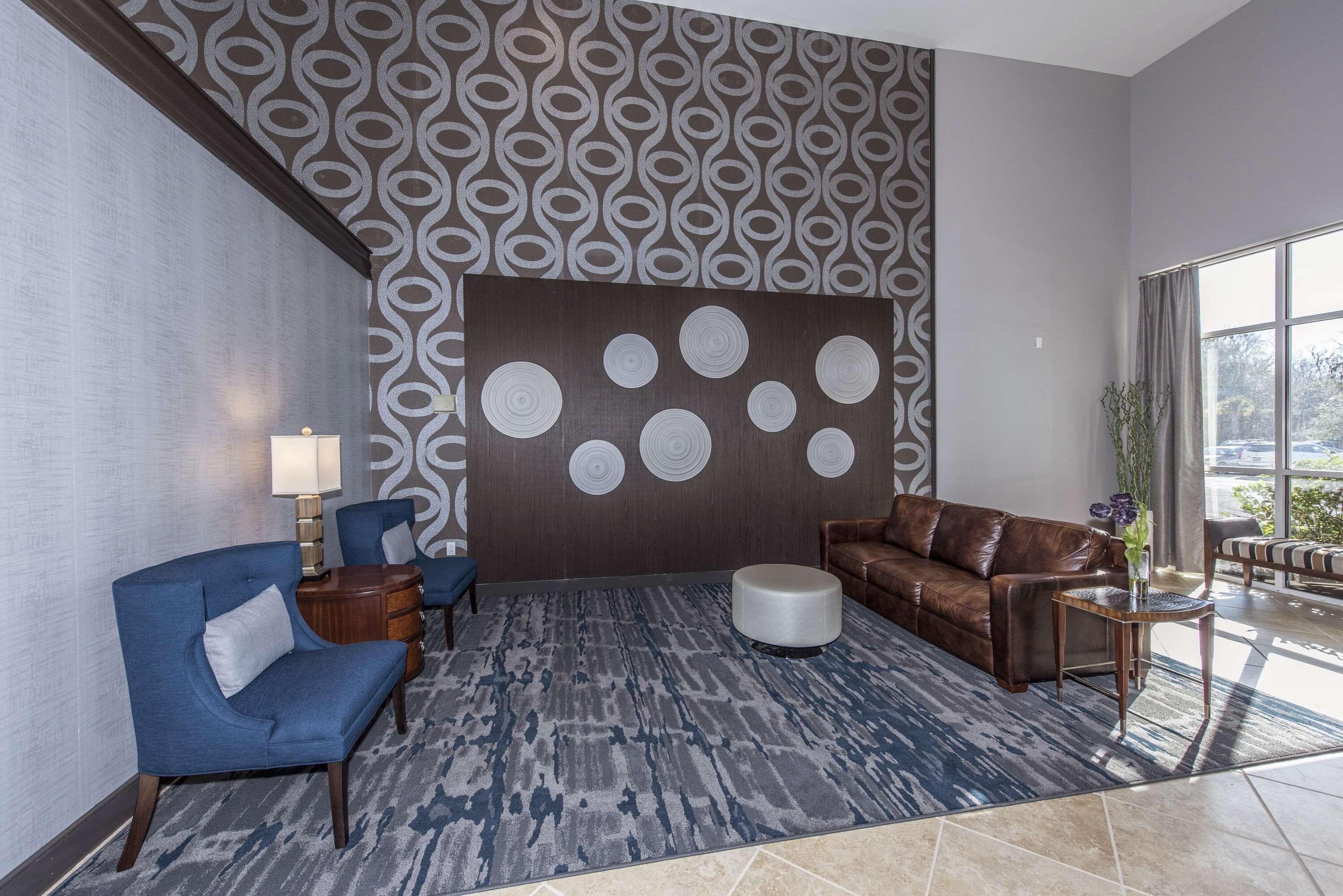A lobby with a patterned wall, a brown leather sofa, a blue armchair, a round ottoman, and a small side table with a vase.