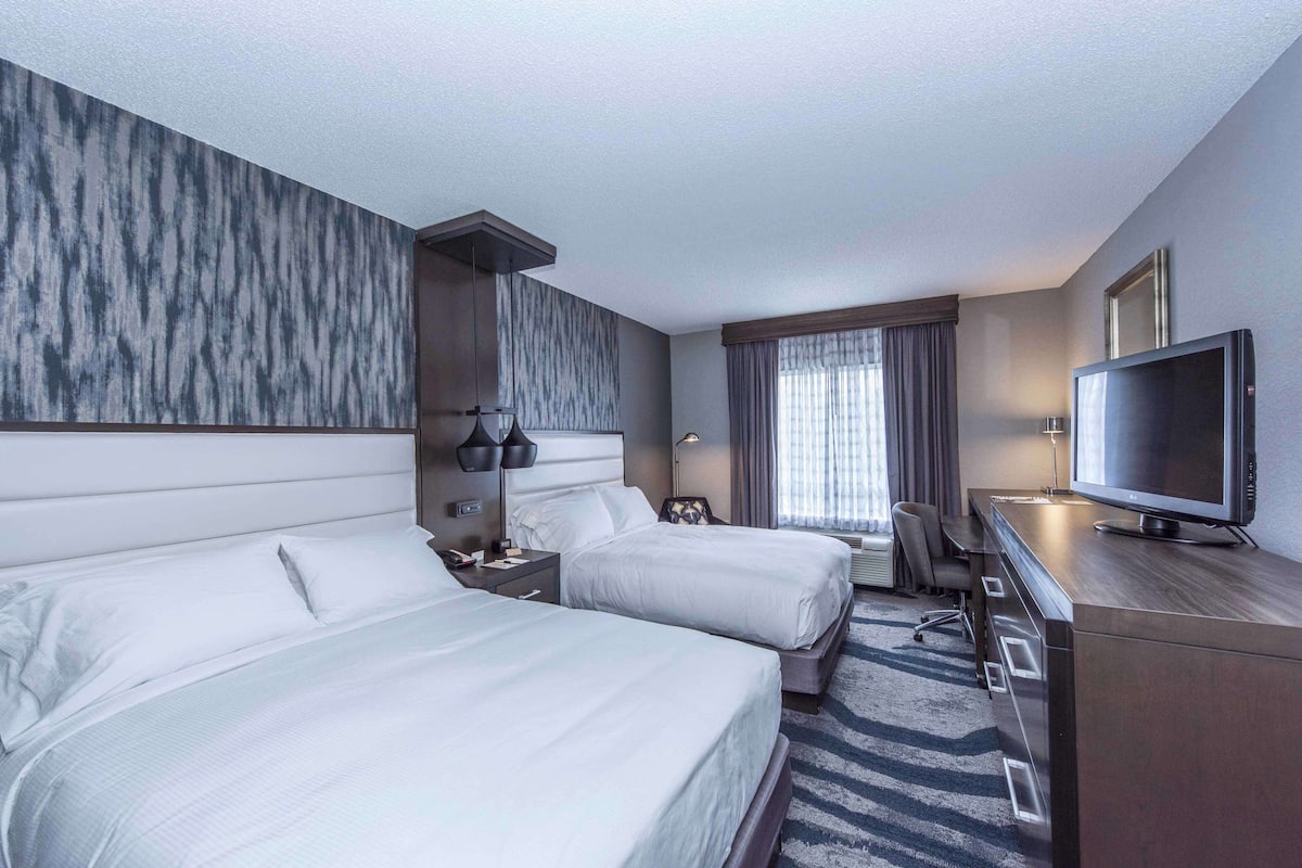 A hotel room with two beds, a desk with a TV, a chair, and a window with curtains.