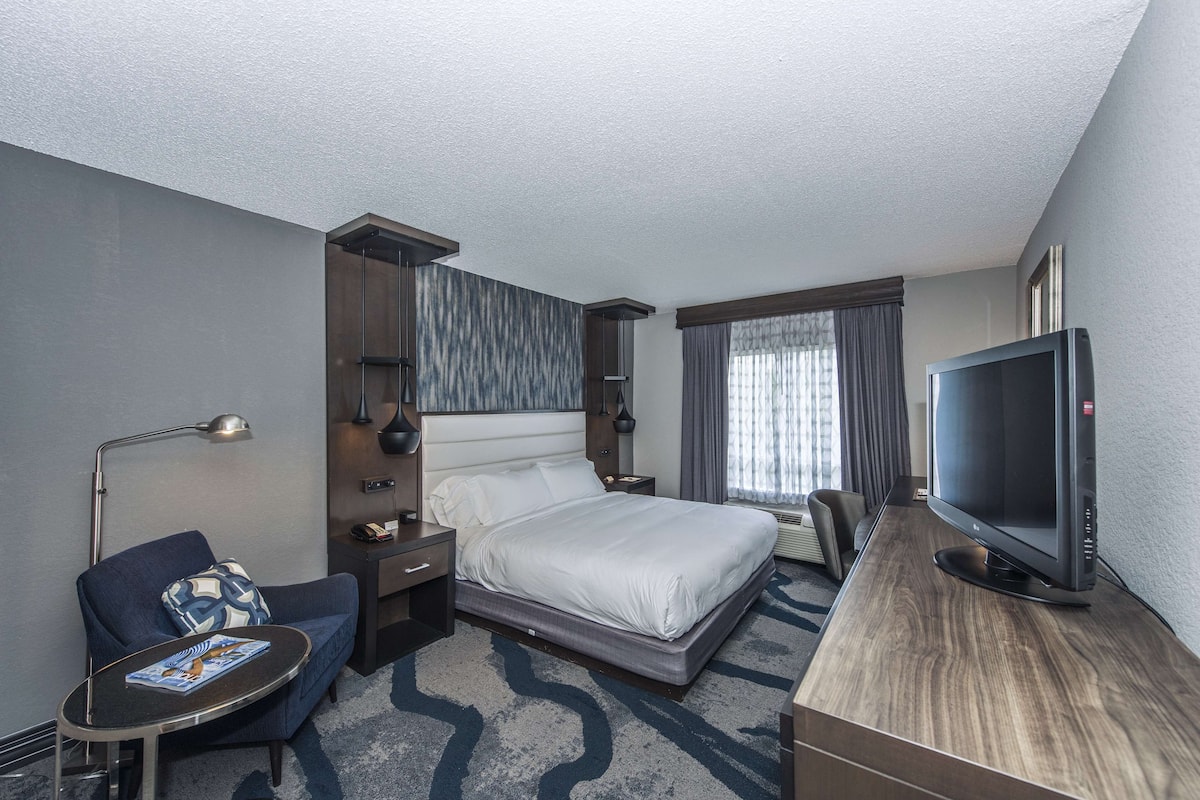 A hotel room with a bed, a desk, a chair, a TV, and a lamp.