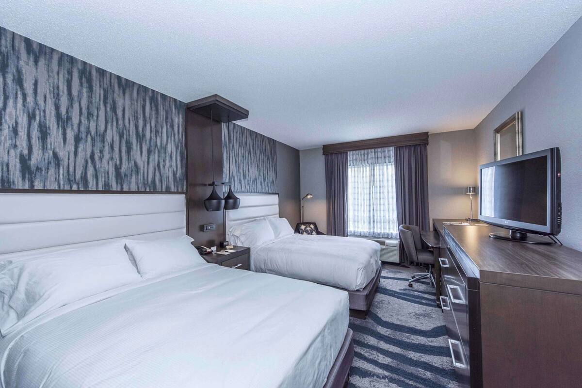 A hotel room with two beds, a desk, a TV, and a large window with curtains.