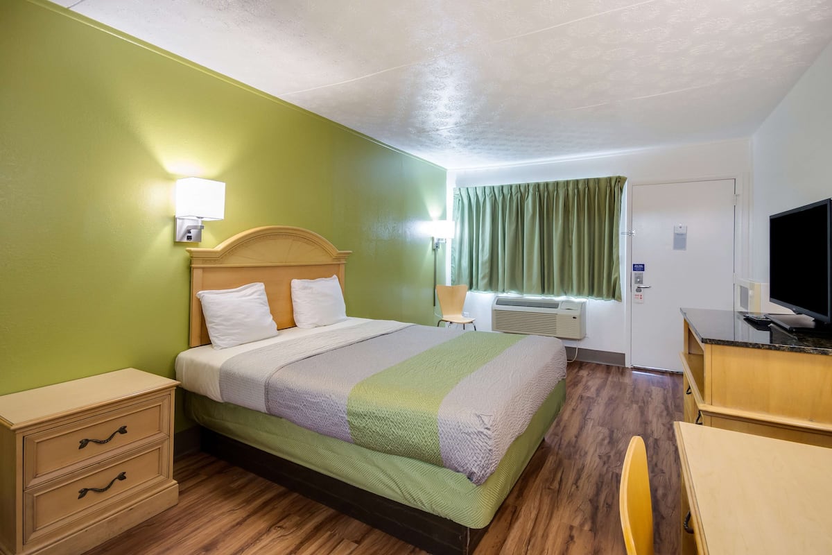 A hotel room with a bed, bedside lamp, a desk with a TV, a chair, a wardrobe, and a window with curtains.