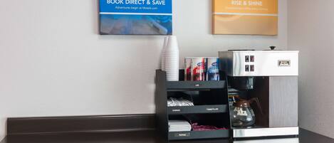 A hotel reception area with a coffee machine, a stack of white cups, and promotional posters on the wall.