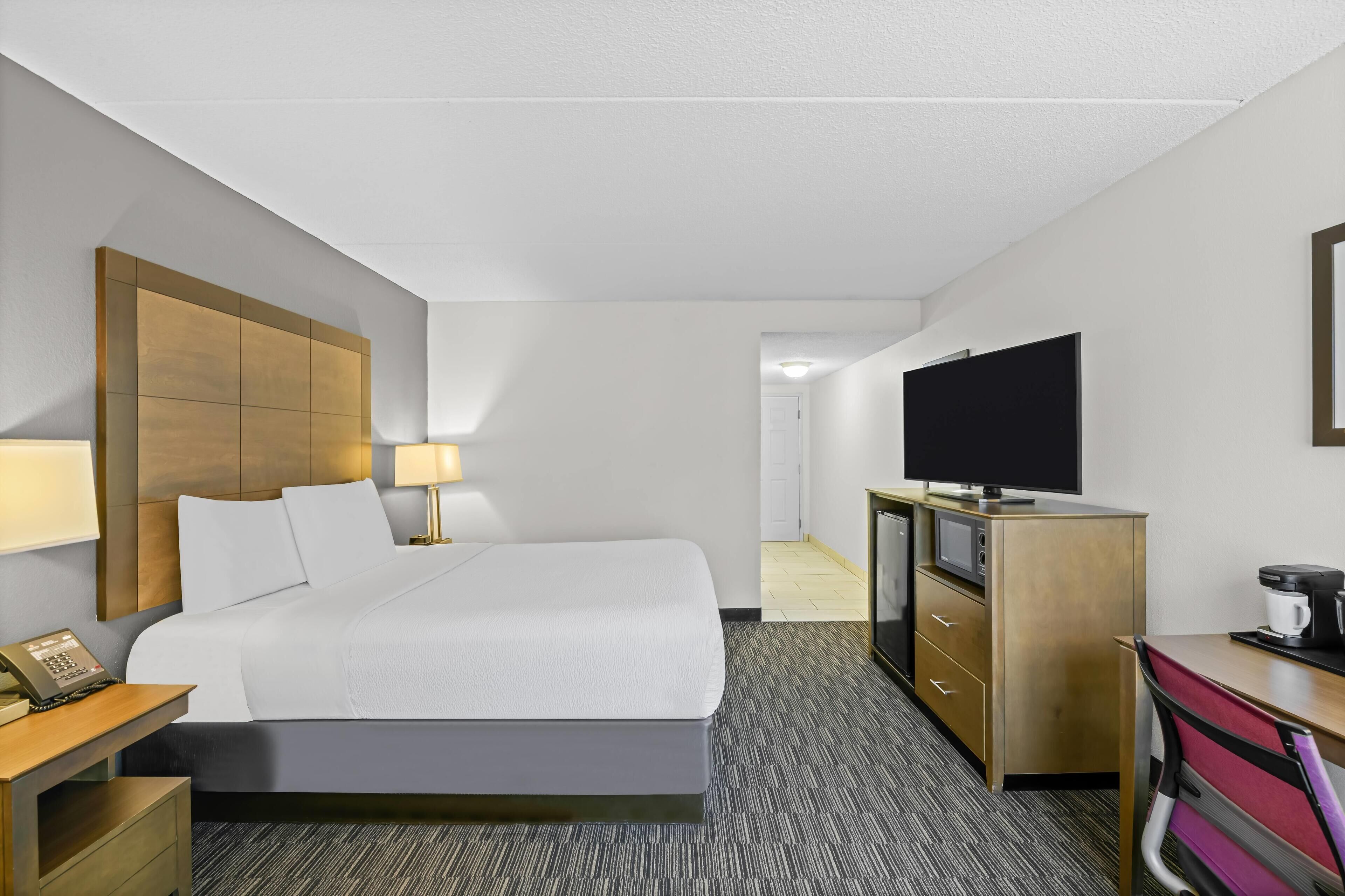 A hotel room with a bed, bedside table, lamp, TV, desk, and a coffee maker.