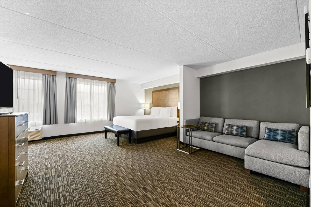 Habitación de hotel con cama, sofá, escritorio y un ventanal con cortinas.