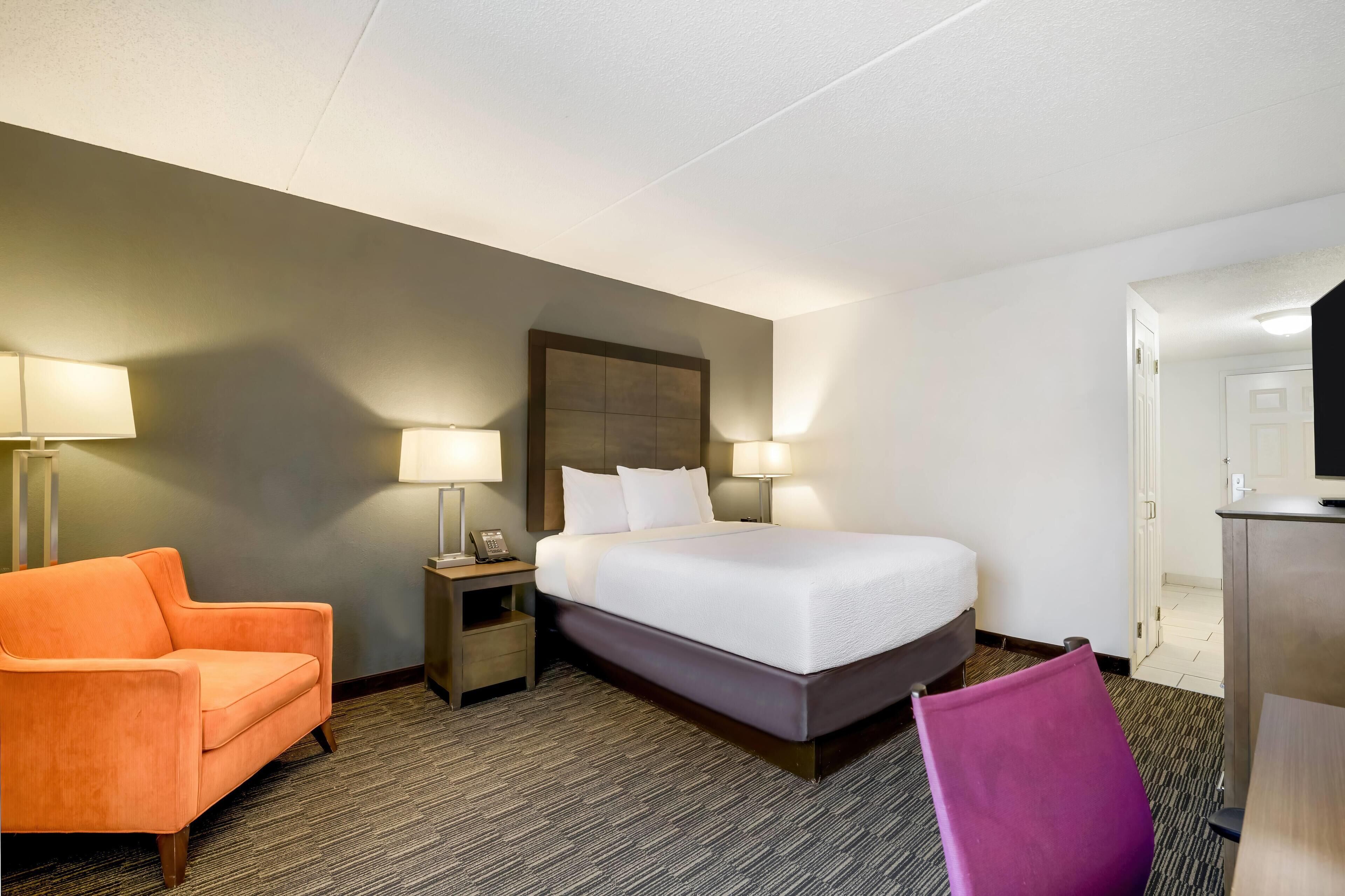 A hotel room with a bed, a nightstand, a lamp, an orange chair, and a desk.