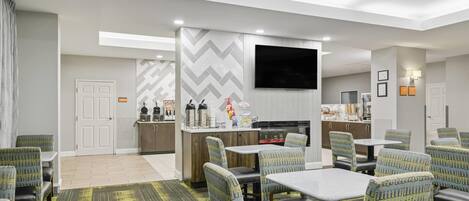 A modern hotel room with a dining area, a kitchenette, and a flat-screen TV mounted on the wall.