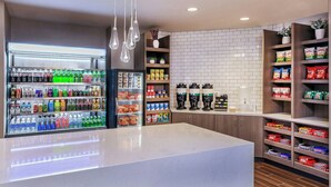 Una kitchenette moderna con un refrigerador lleno de bebidas, una cafetera y estantes con bocadillos.