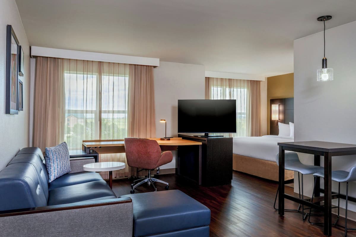 A modern hotel room with a sofa, a desk with a chair, a TV, a bed, and a small table with chairs.