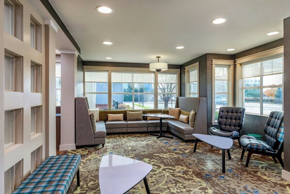 Uma área de estar moderna com sofá seccional, uma mesa pequena e duas poltronas. Janelas amplas oferecem luz natural e vista para o exterior.