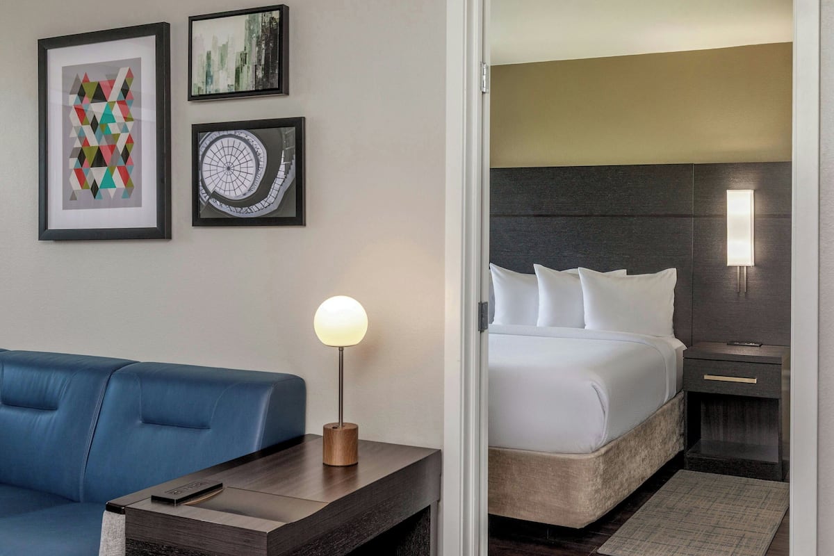 A hotel room with a bed, a blue sofa, a bedside table with a lamp, and framed artwork on the wall.