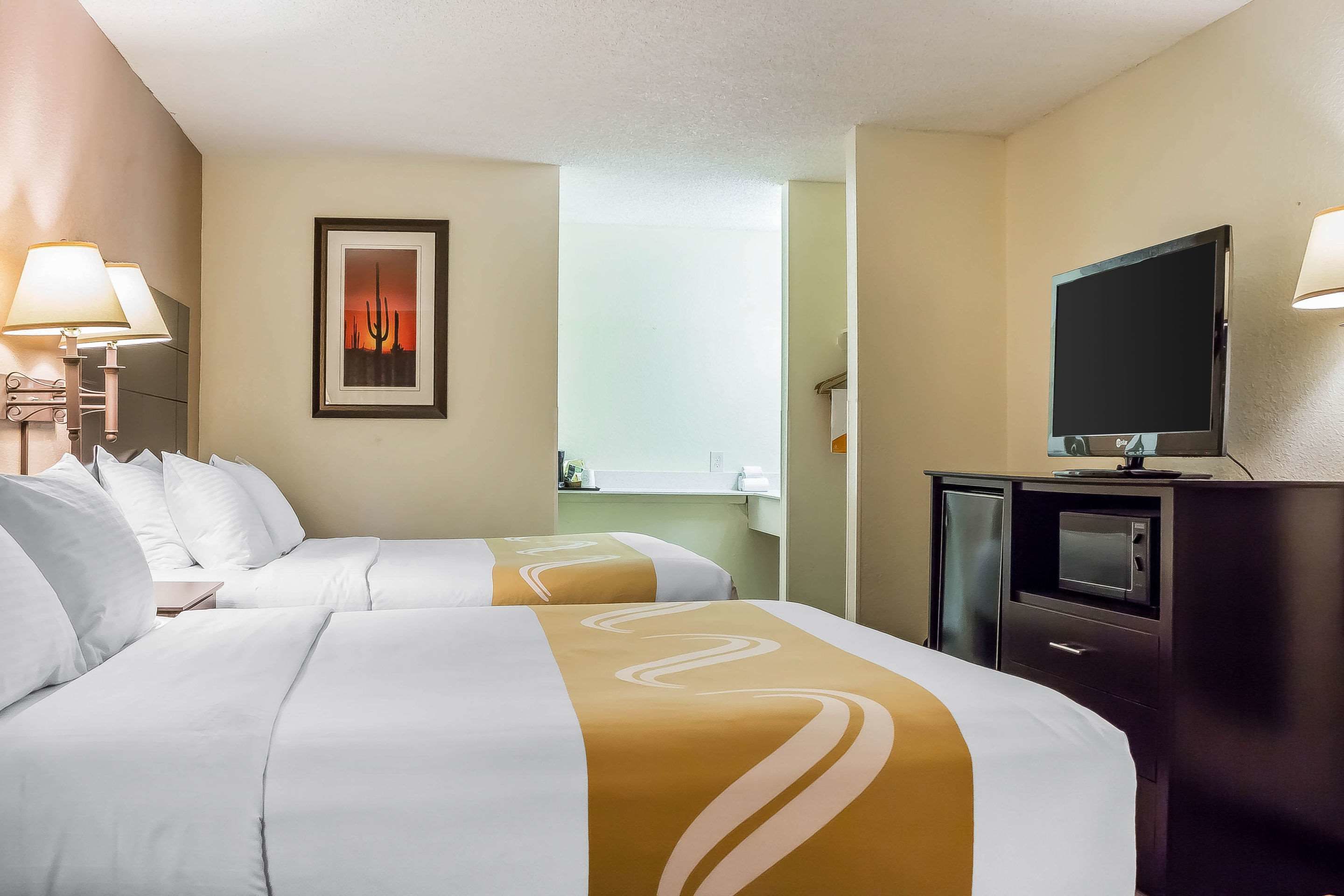 Quarto de hotel com duas camas, uma televisão, uma escrivaninha e um quadro na parede.