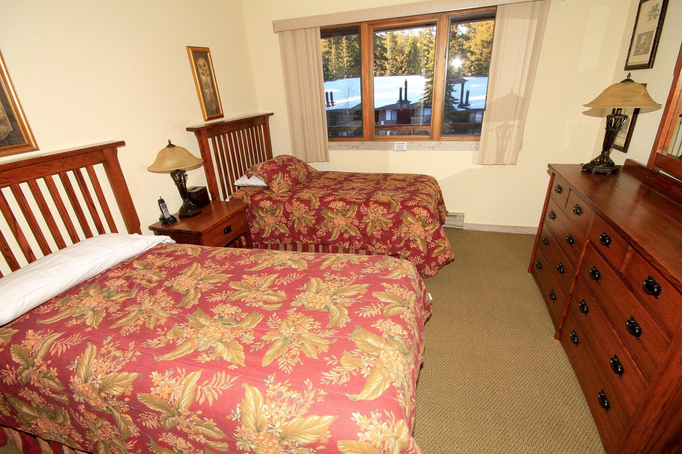 A bedroom with two beds, wooden headboards, a dresser, and a window with a view of trees and a cabin.
