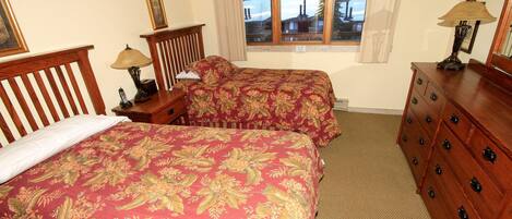 A bedroom with two beds, wooden headboards, a dresser, and a window with a view of trees and a cabin.