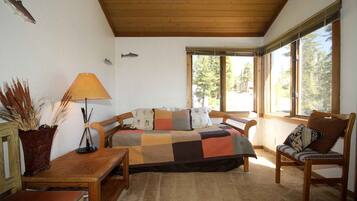 A cozy room with a wooden ceiling, a large window with a view of trees, a bed with a checkered blanket, a wooden table, and a lamp.