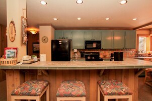 A kitchen with wooden cabinets, a refrigerator, a microwave, and a bar area with stools.