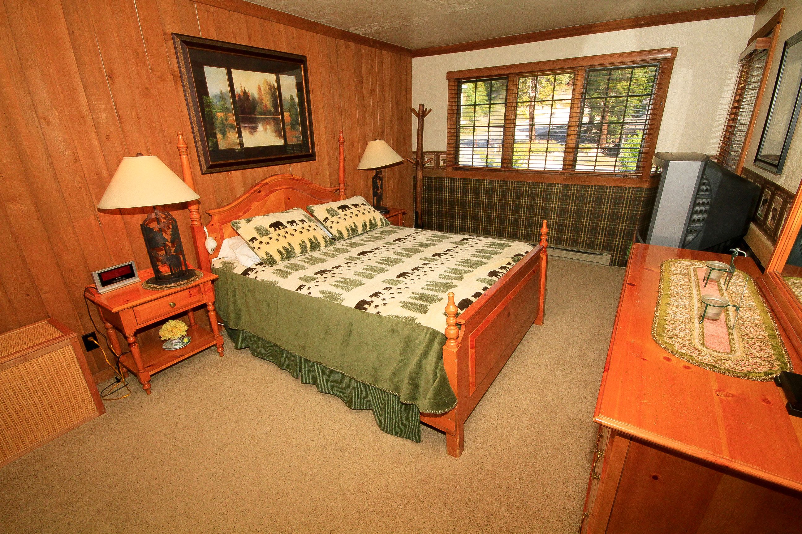 A bedroom with a wooden bed, two bedside tables with lamps, a dresser, a radiator, and a window with blinds.