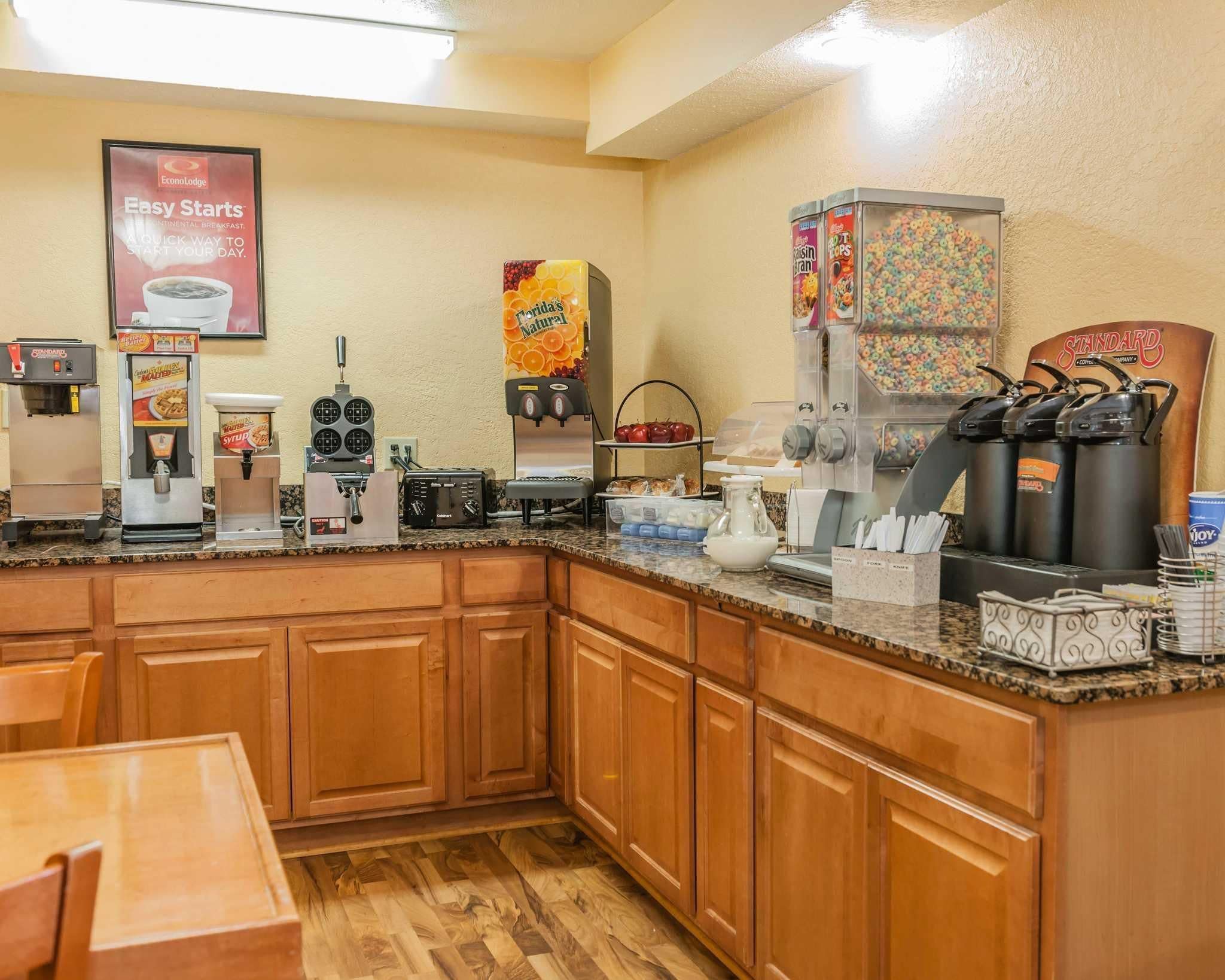Un área de desayuno de hotel con una cafetera, dispensador de jugo, dispensador de cereal y una jarra de café.
