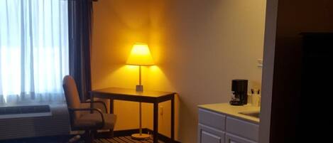 Quarto de hotel com cama, escrivaninha, cadeira, luminária, cafeteira e janela com cortinas.