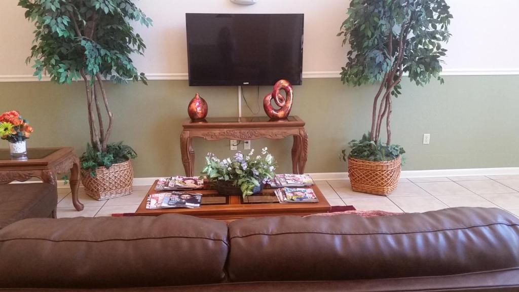 A living room with a brown leather sofa, a wooden coffee table with magazines, two potted plants, and a flat-screen TV mounted on the wall.