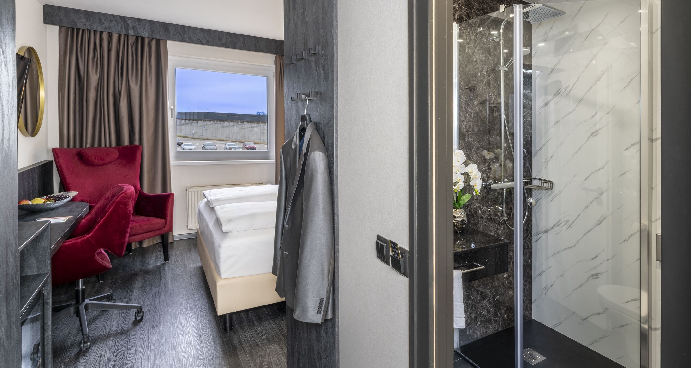Habitación de hotel con cama, una silla roja, escritorio, ducha y vista al exterior.