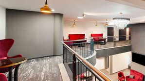 A modern hotel lobby with a mezzanine level, red seating, and a chandelier. - PLAZA INN Mühldorf am Inn (Muhldorf am Inn)