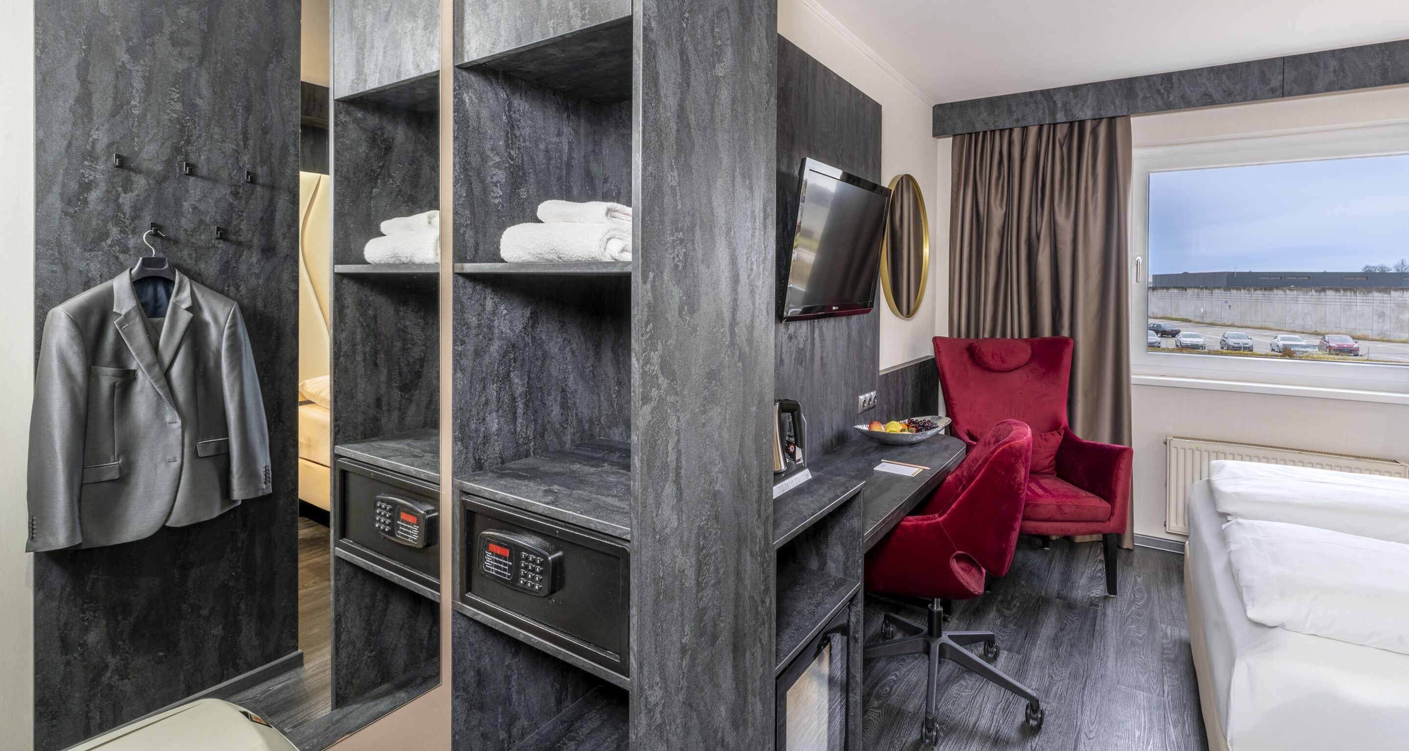 A hotel room with a bed, a desk with a chair, a TV, a red chair, and a wardrobe with shelves.