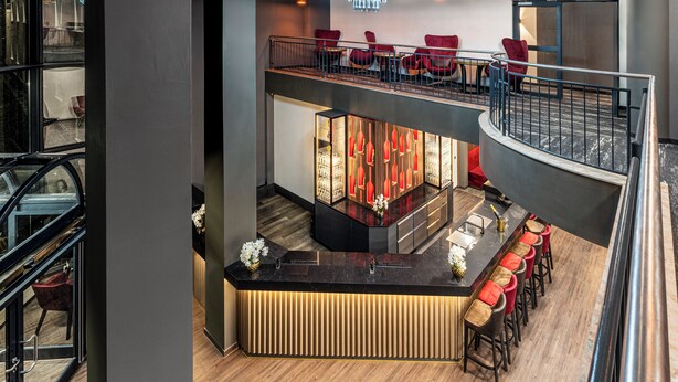 A modern hotel lobby with a central bar area, seating on the upper level, and a staircase connecting floors. - PLAZA INN Mühldorf am Inn (Muhldorf am Inn)