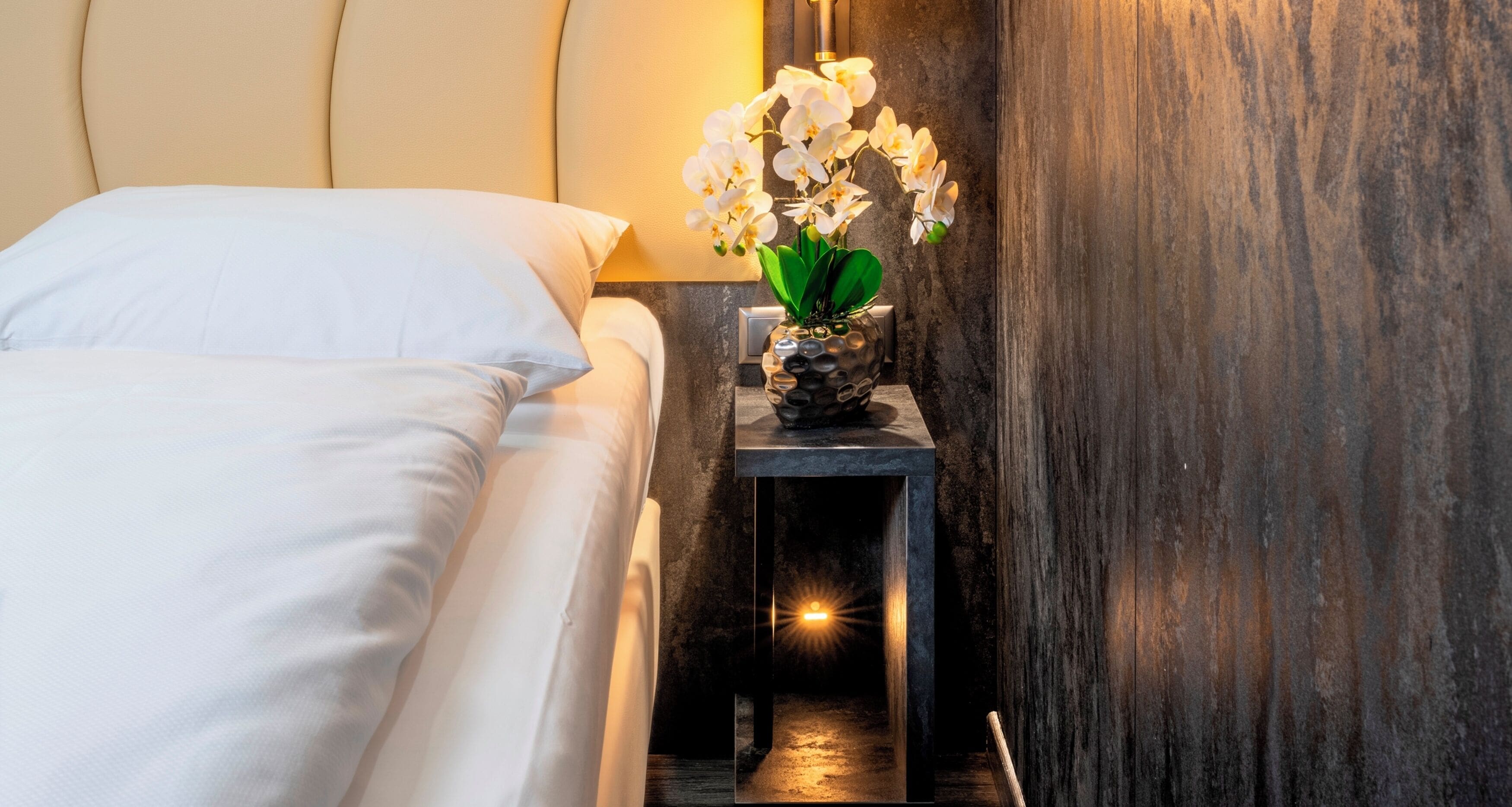 Una cama bien hecha con sábanas blancas, una mesita de noche con un jarrón de orquídeas blancas y una lámpara de pared.