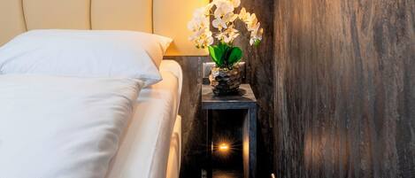 Una cama bien hecha con sábanas blancas, una mesita de noche con un jarrón de orquídeas blancas y una lámpara de pared.