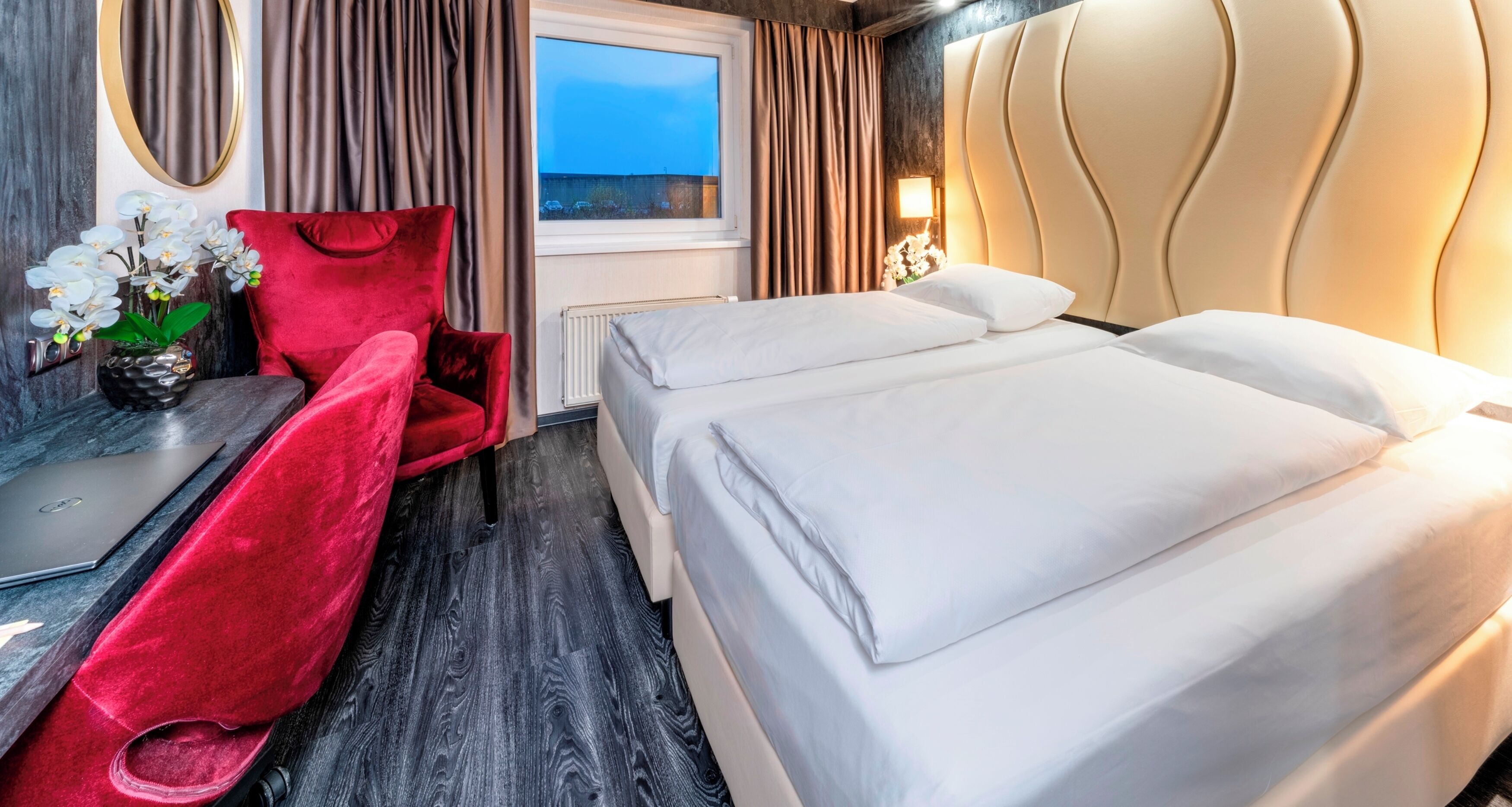 Een hotelkamer met een bed, een bureau met een laptop, een rode stoel, een vaas met bloemen en uitzicht op de zee.