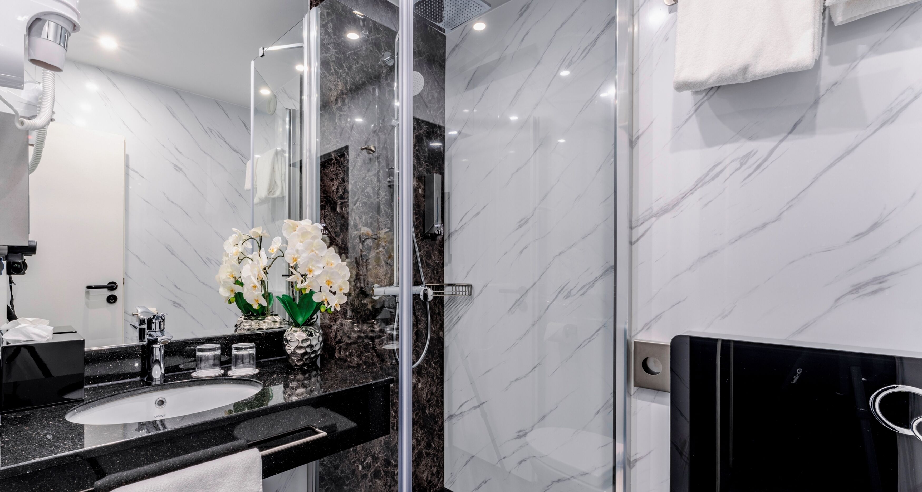A modern bathroom with a black marble sink, a glass shower enclosure, and white marble walls.