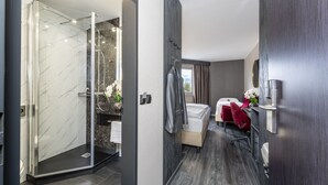 A modern hotel room with a glass-enclosed shower, a vanity with a marble countertop, a bed with a red chair, and a view of the outdoors through a window. - PLAZA INN Mühldorf am Inn (Muhldorf am Inn)