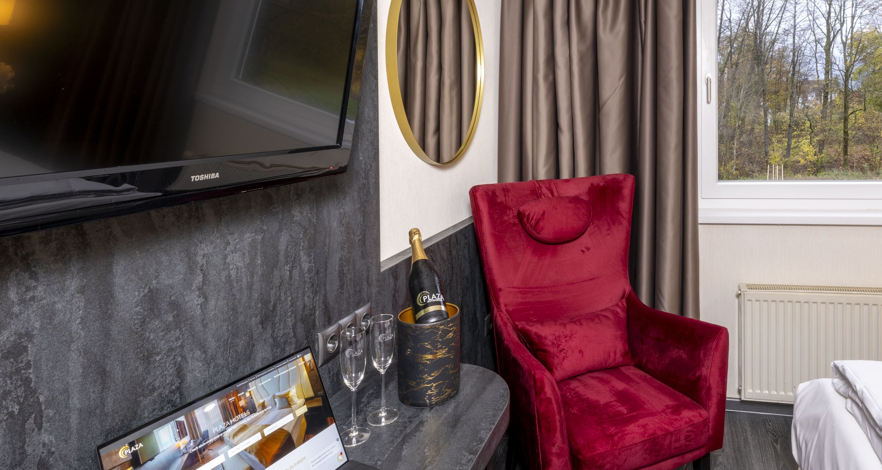 Habitación de hotel moderna con una televisión de pantalla plana, un sillón de terciopelo rojo, un minibar con champán y vistas a árboles por la ventana.