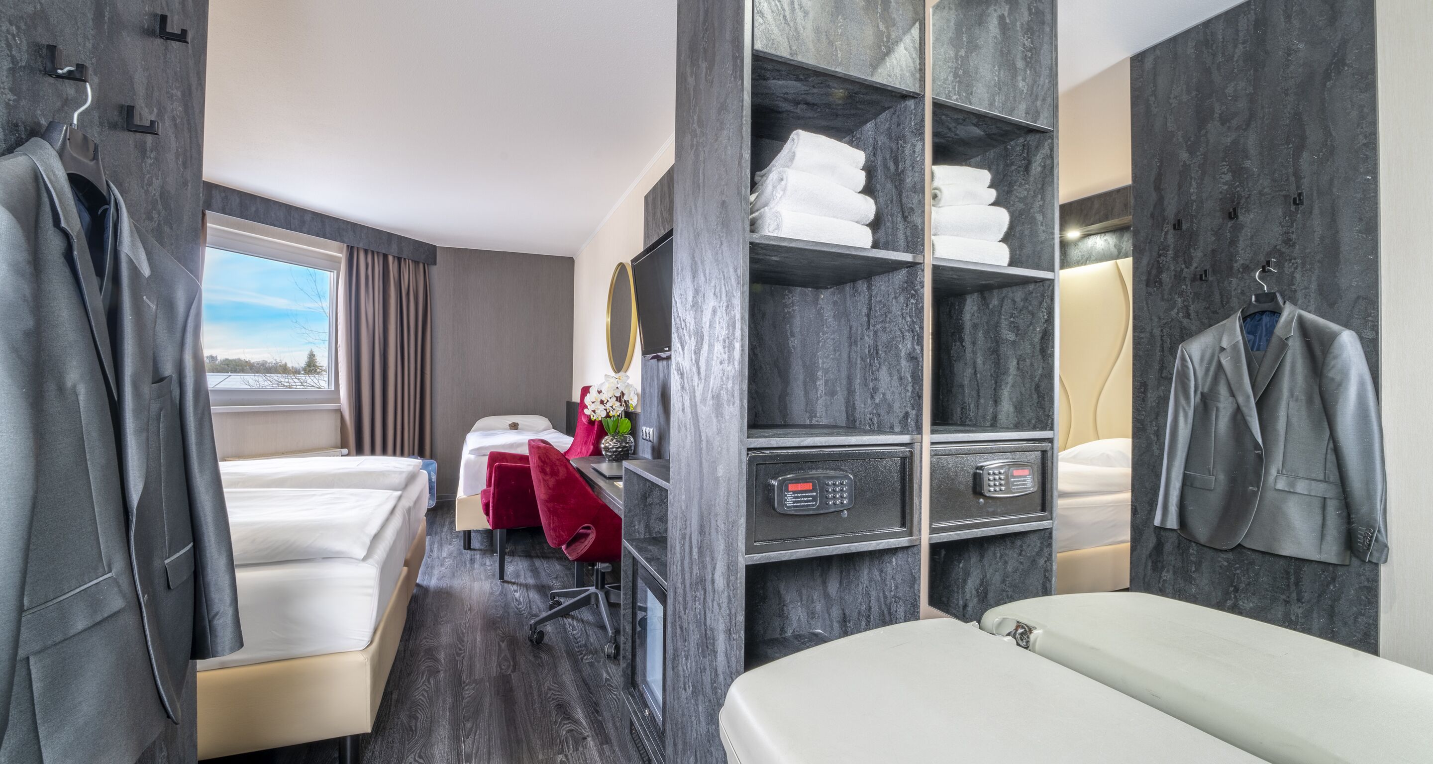 A hotel room with a bed, a desk, a chair, a vanity with a mirror, a robe, and a coat rack.