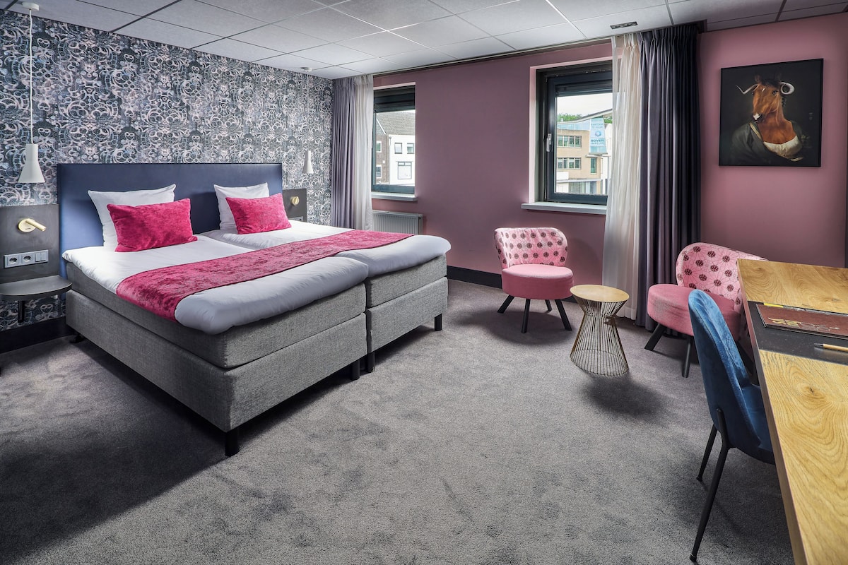 A modern hotel room with a grey bed, pink pillows, a small round table, and a chair.