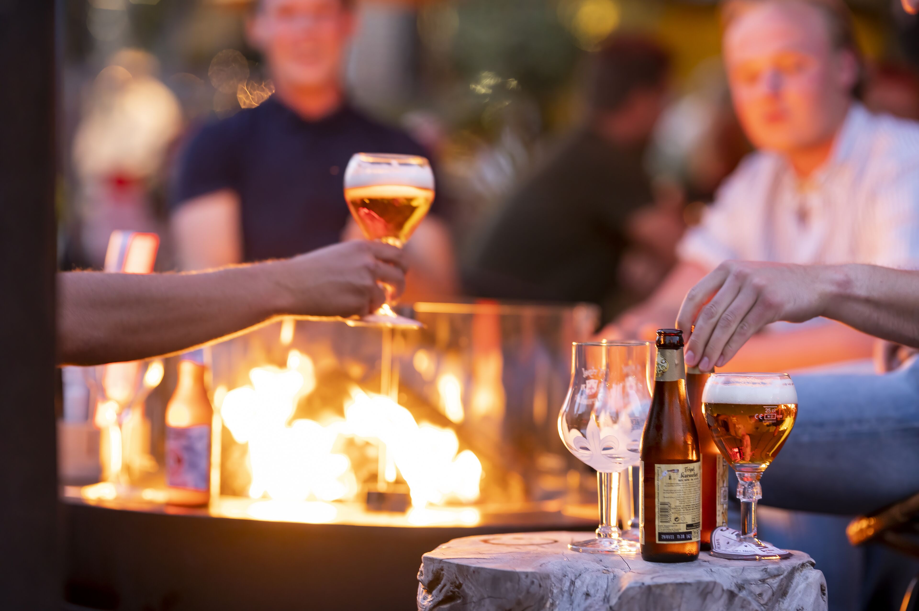A table with a bottle and glasses, a fire in the background, and people in the distance.