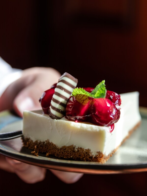 Una porción de pastel de queso con base desmenuzable, coronada con una cereza y una galleta cubierta de chocolate.