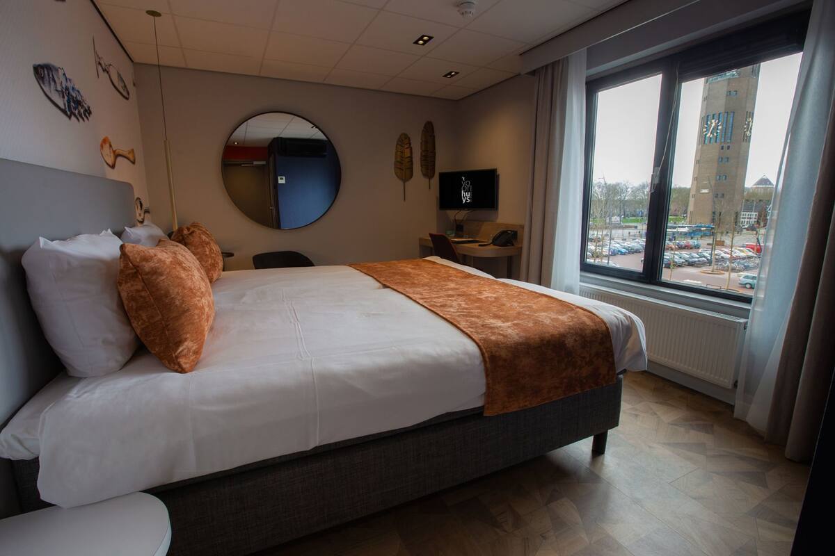 A hotel room with a bed, a desk, a chair, a mirror, and a window with a view of a parking lot and buildings.